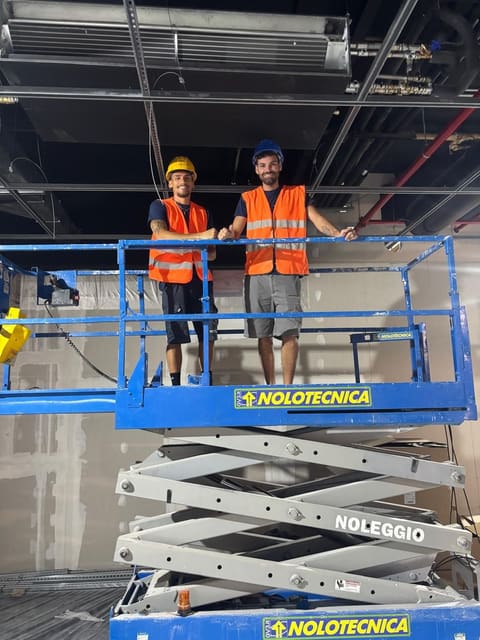 Due tecnici con casco e gilet arancione ad alta visibilità in piedi su una piattaforma elevabile, sotto un soffitto con tubazioni e impianti a vista.